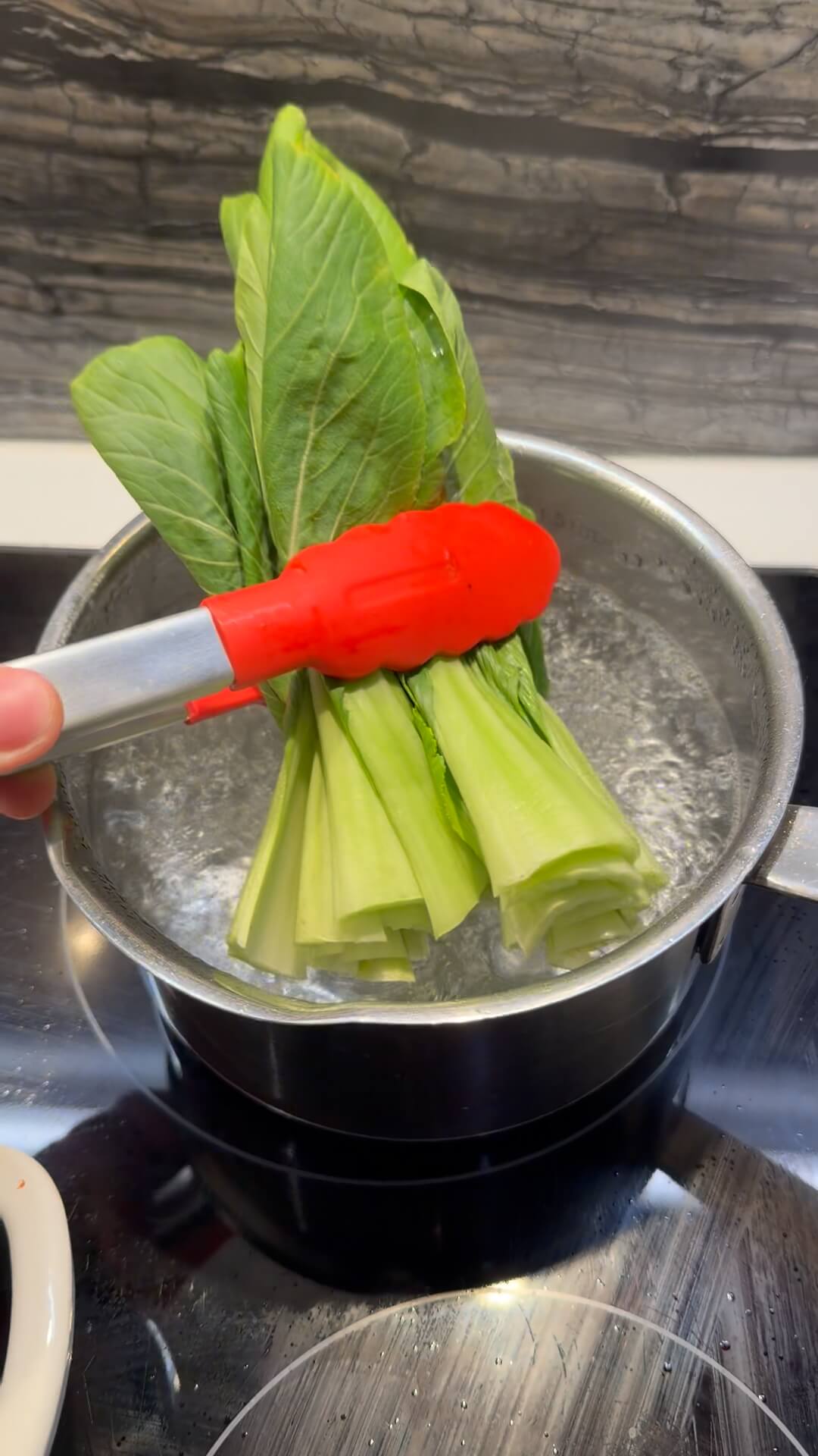 Fresh green bok choy being placed into a pot of boiling water with red tongs.