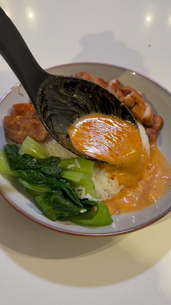 A ladle pouring creamy Thai red curry sauce over seared chicken thighs, bok choy, and jasmine rice in a bowl.