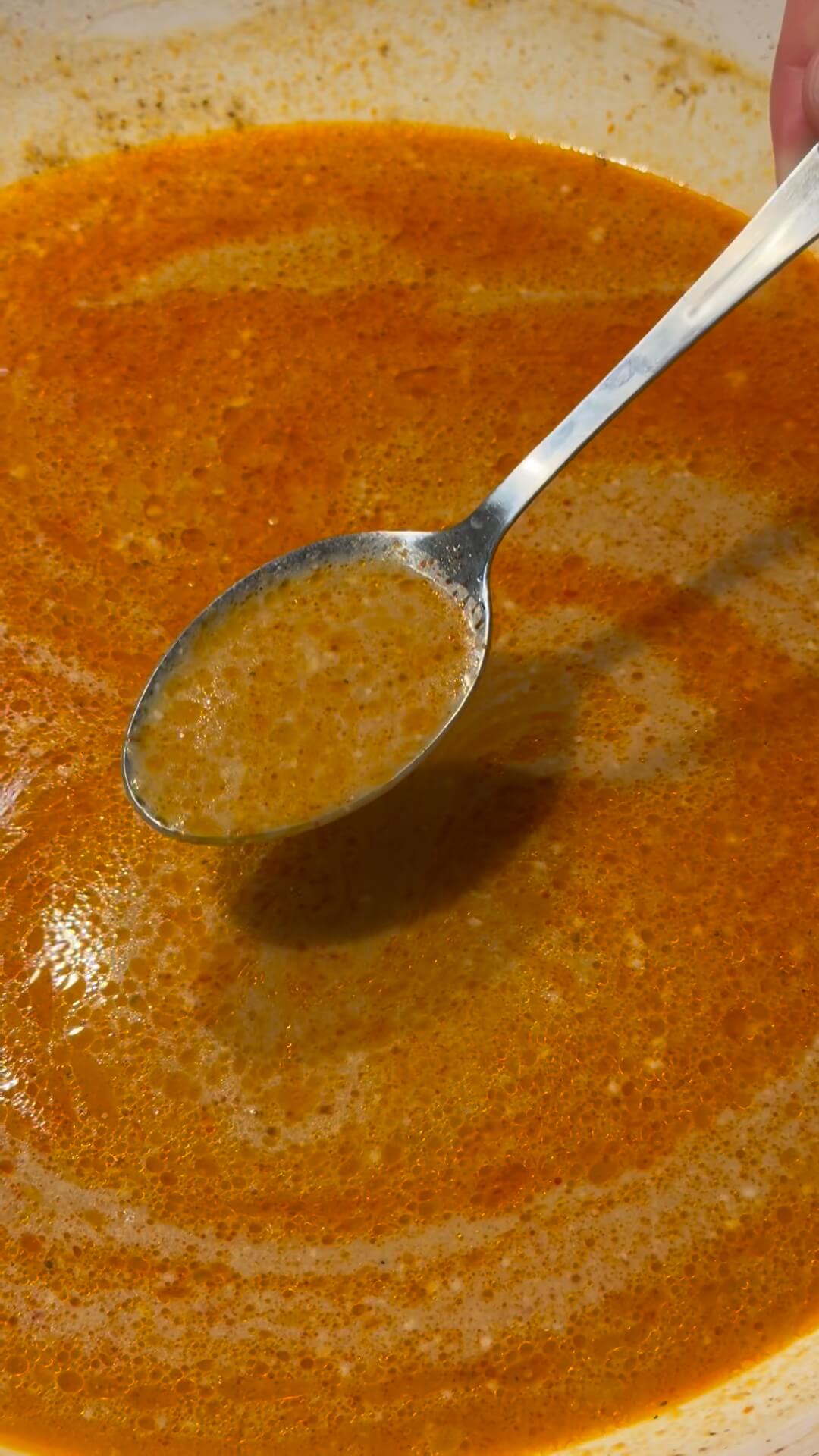 A metal spoon stirring a vibrant, creamy orange-red Thai curry sauce in a pan.