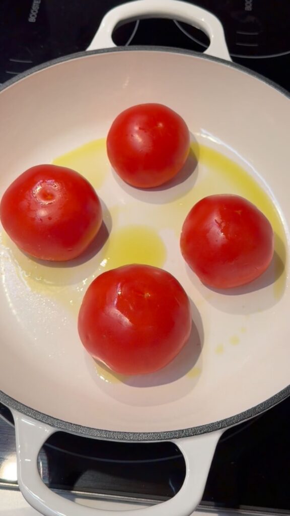 Four whole Roma tomatoes placed upside down in a white skillet with olive oil, ready to be steamed and peeled.