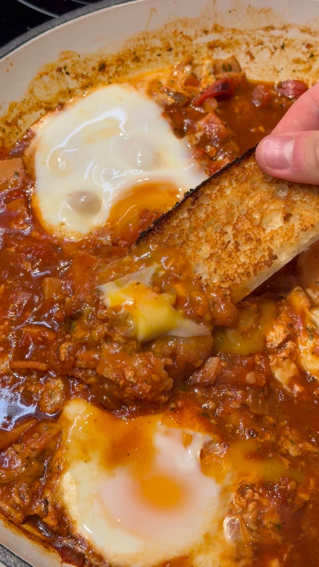 A close-up shot of homemade Shakshuka (Middle Eastern Baked Eggs) in a pan with runny poached eggs, being scooped up by a thick slice of toasted sourdough bread.