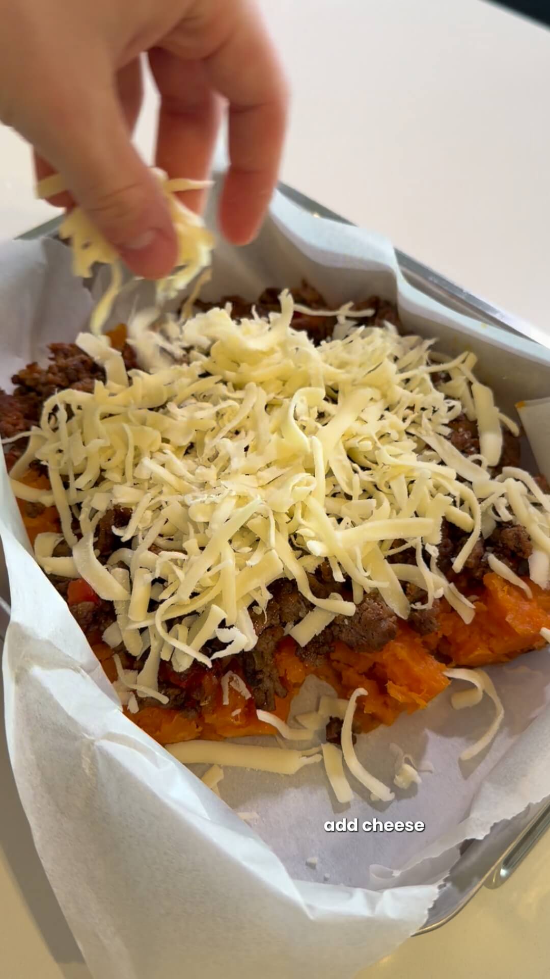 A hand sprinkling shredded mozzarella cheese over a layer of cooked beef mince and mashed sweet potato.