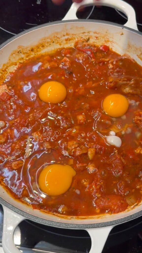 Three raw eggs cracked into "wells" within a simmering, chunky tomato and bacon shakshuka base in a white pan.
