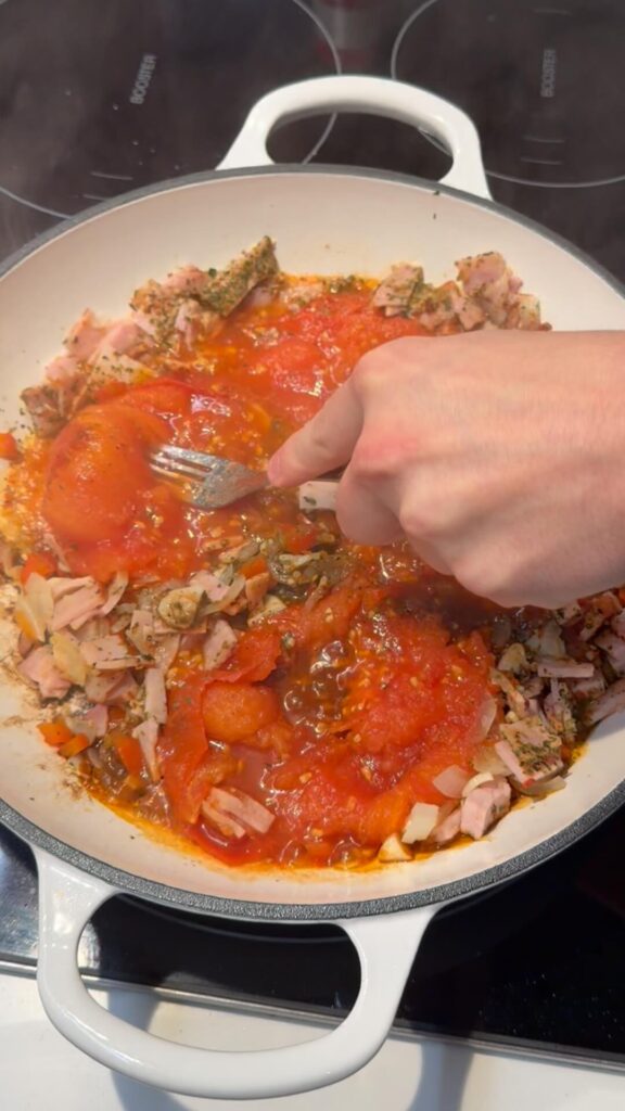 A hand using a fork to mash softened, peeled whole tomatoes directly into a pan with sautéed vegetables.