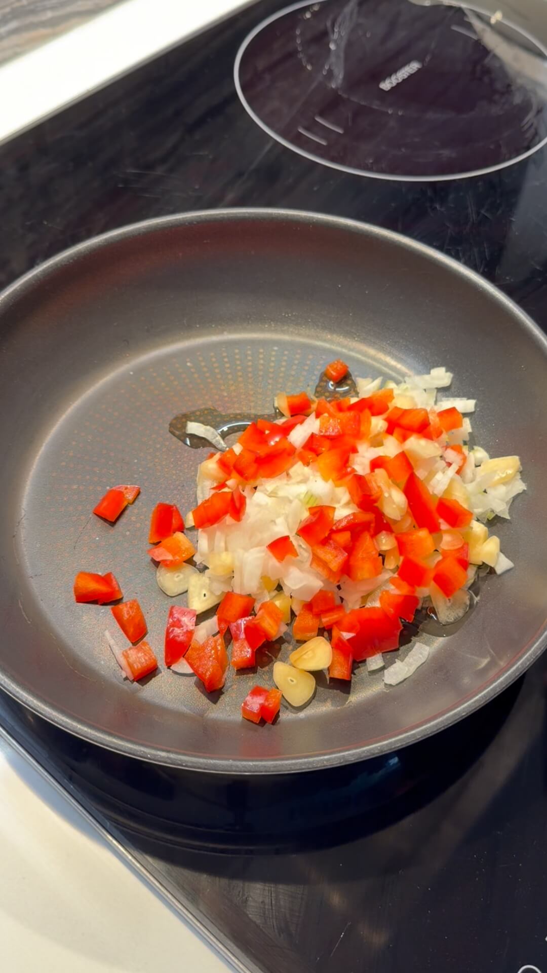 Diced white onion and red capsicum cooking in a grey non-stick frying pan.