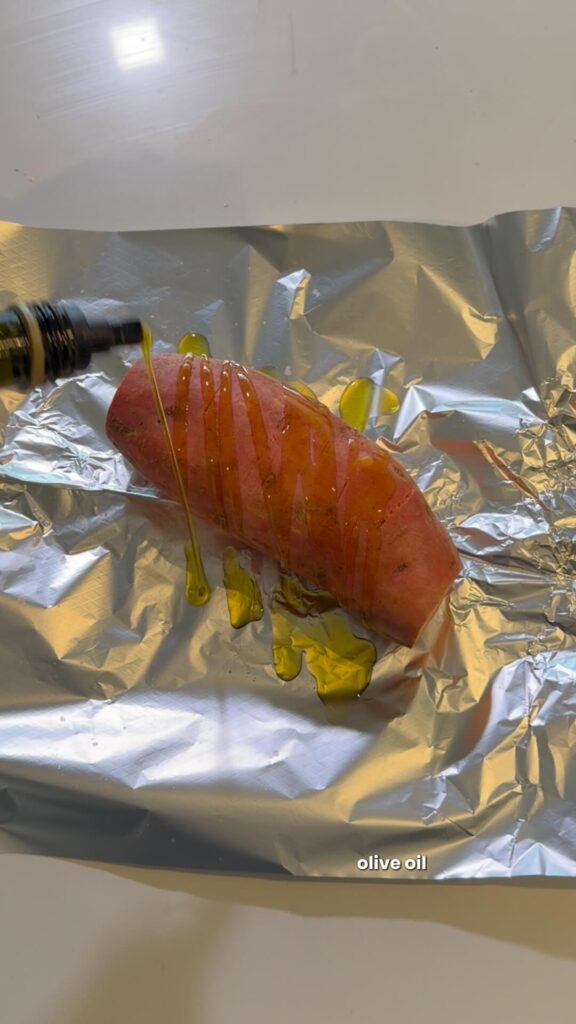 A whole sweet potato being drizzled with olive oil on a sheet of aluminum foil.