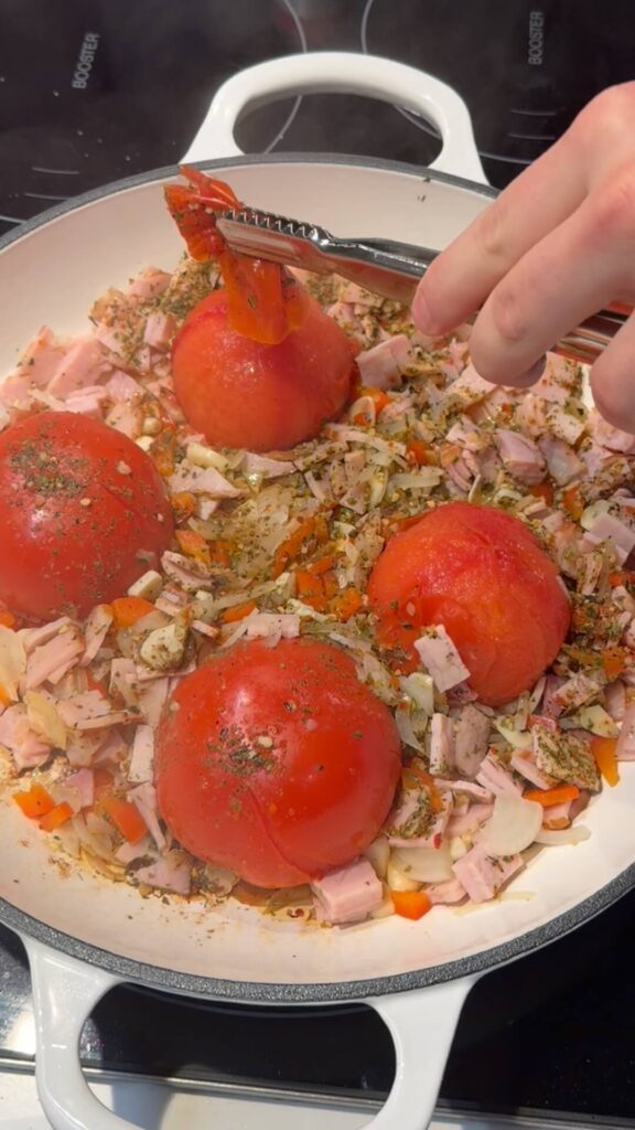 Using tongs to peel the skin off a whole steamed tomato in a pan filled with sautéed onions and bacon.