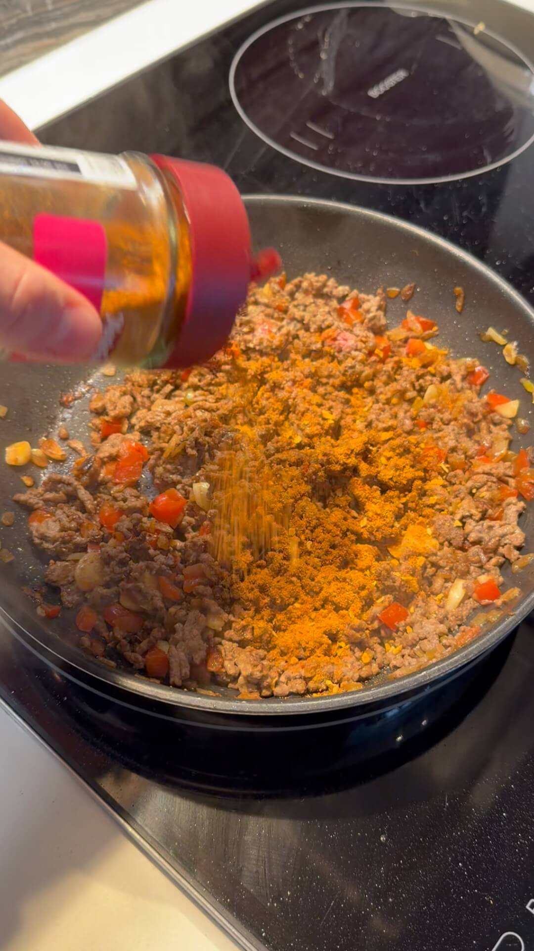 A hand shaking orange taco seasoning from a glass jar into a pan of browned beef mince, onions, and capsicum.